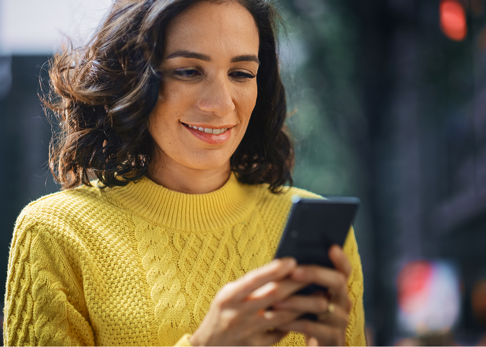 Woman uses phone outdoors. She's wearing a yellow sweater.