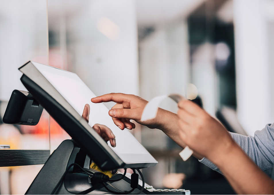 Hands operate a touchscreen POS system, dispensing a receipt; retail environment.