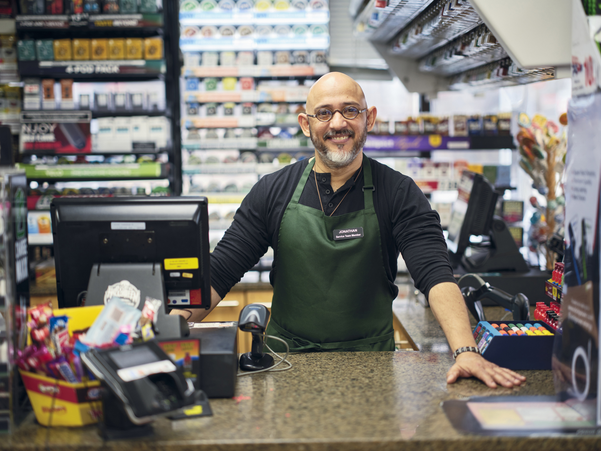 Clerk smiles, leaning on counter, in convenience store. JONATHAN Service Team Member